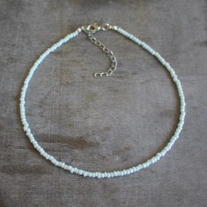 Op de afbeelding: Een delicate, lichtblauwe kralenketting met een zilverkleurige sluiting en verlengketting. De ketting bestaat uit kleine, gelijkmatig verdeelde kralen, wat een minimalistisch ontwerp creëert. De ketting is ongeveer 35,5 cm lang, plus een verlenging van 5 cm.