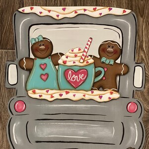 May include: A gray truck-shaped wooden decoration featuring gingerbread people and a mug with the word "love." The gingerbread couple is holding a mug with whipped cream and a candy cane straw. The truck has the text "XOXO" on the license plate.