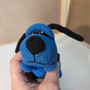 Può includere: Un piccolo giocattolo di peluche a forma di cane, blu con orecchie nere. Il cane ha un grande naso nero e un'espressione amichevole. Il giocattolo è tenuto in mano, suggerendo le sue piccole dimensioni.