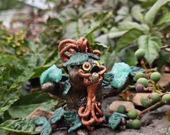 Puede incluir: Una escultura artesanal de una criatura con forma de árbol, con tronco marrón, hojas verdes y detalles turquesa. La criatura tiene ojos grandes y expresivos y una expresión juguetona. La escultura está sobre un fondo natural de follaje verde y piedra.