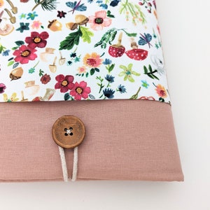May include: A fabric item with a floral pattern featuring red, pink, and yellow flowers, mushrooms, and acorns on a white background. The bottom section is a solid dusty rose color. A wooden button with a white cord closure is visible.