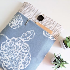 May include: A blue fabric laptop sleeve with a white floral pattern. The sleeve has a pocket on the outside with a brown fabric lining and a button closure.