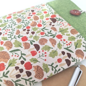 May include: A green fabric iPad case with a brown button closure. The front of the case features a beige fabric with a repeating pattern of hedgehogs, apples, leaves, and mushrooms.