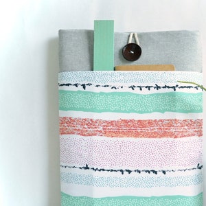 May include: A gray and white striped fabric laptop sleeve with a green and white patterned pocket. The sleeve is sitting on a white surface with a vase of yellow daffodils.