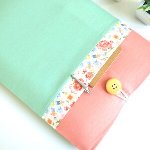 May include: A mint green laptop sleeve with a floral patterned pocket and a pink flap. The flap has a yellow button and a white string closure.