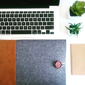 May include: A gray laptop sleeve with a brown leather flap and a red button closure. The sleeve is made of a textured fabric and has a subtle speckled pattern.