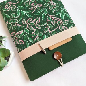 May include: A green laptop sleeve with a pink and green floral pattern. The sleeve has a beige flap with a silver pen and a brown button.