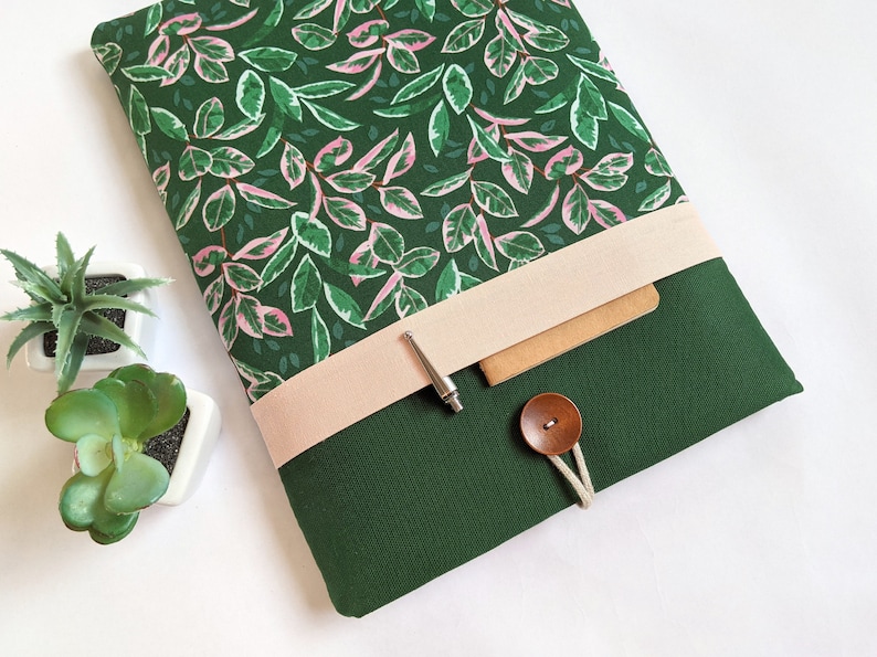 May include: A green laptop sleeve with a pink and green floral pattern. The sleeve has a beige flap with a silver pen and a brown button.