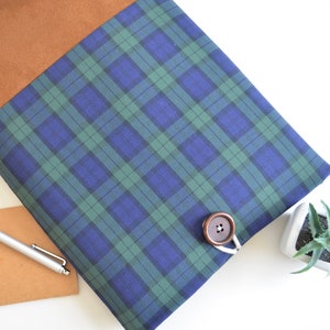 May include: A blue and green plaid laptop sleeve with a brown suede flap and a brown button closure.
