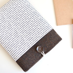 May include: A brown and white book sleeve with a button closure. The sleeve is made of a fabric with a tweed-like texture. The front of the sleeve is covered in a white fabric with black text.