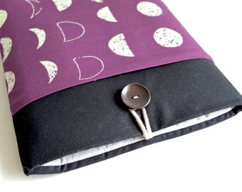 Puede incluir: Una funda para portátil de color morado con un patrón de lunas y círculos blancos. La funda tiene una solapa negra con un botón marrón y un cierre de cuerda blanca.