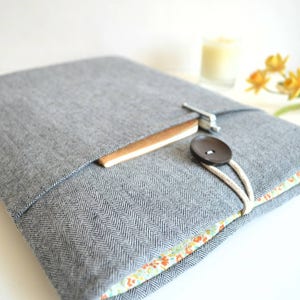 Puede incluir: Una funda para portátil gris con un patrón de espiga y un forro floral. La funda tiene una solapa con cierre de botón y un bolsillo para un bolígrafo o un lápiz.