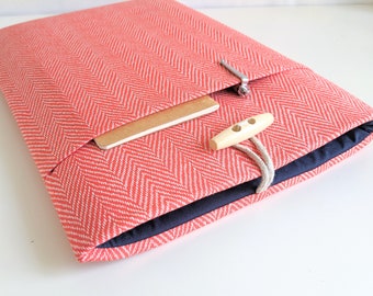 May include: A coral pink laptop sleeve with a herringbone pattern. The sleeve has a pocket on the front and a wooden toggle closure.