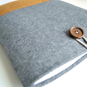 Pode incluir: Uma capa para laptop de linho cinza com fecho de botão castanho e aba de camurça castanha.
