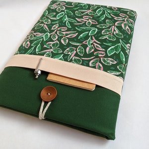 May include: A green laptop sleeve with a pink and green floral pattern. The sleeve has a flap with a beige fabric strap and a brown button closure. A pen and a notebook are tucked into the flap.