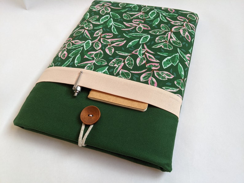 May include: A green laptop sleeve with a pink and green floral pattern. The sleeve has a flap with a beige fabric strap and a brown button closure. A pen and a notebook are tucked into the flap.