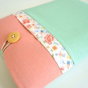 May include: A light pink fabric pouch with a floral patterned trim and a wooden button closure. The pouch is closed and has a light green fabric backing.