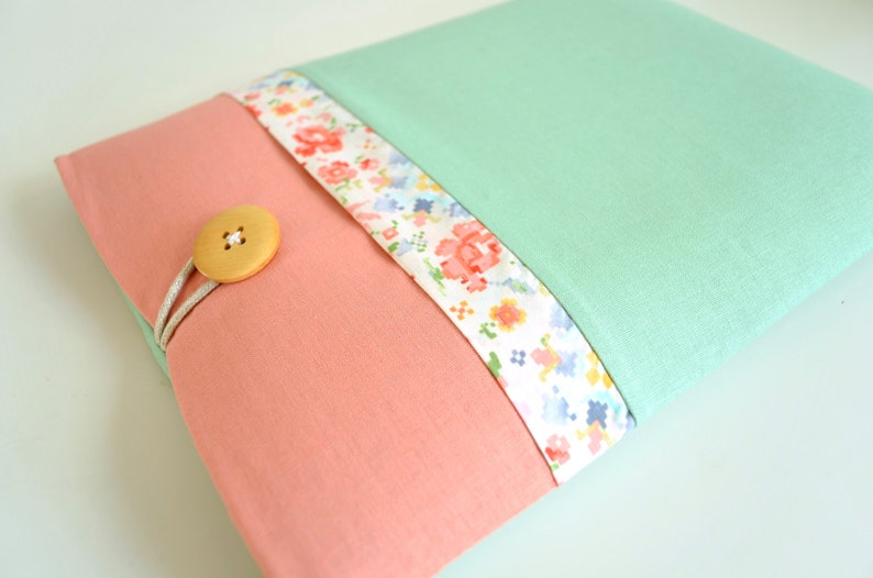 May include: A light pink fabric pouch with a floral patterned trim and a wooden button closure. The pouch is closed and has a light green fabric backing.