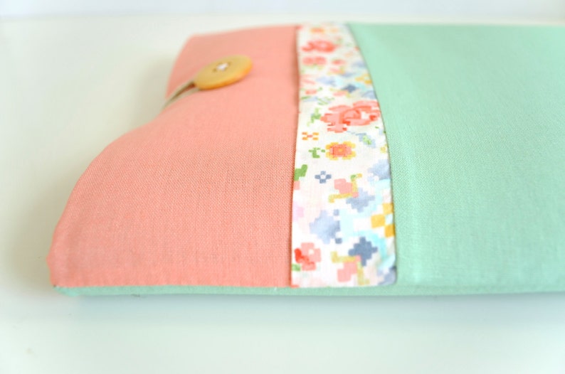 May include: A pink and mint green fabric laptop sleeve with a floral patterned fabric trim and a wooden button.