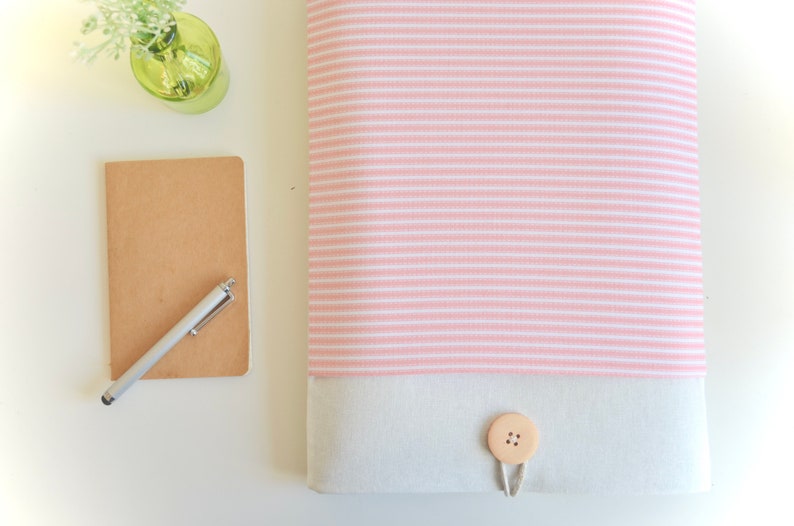 Op de afbeelding: Een roze en wit gestreepte laptophuls met een witte canvas klep en een houten knoopsluiting.
