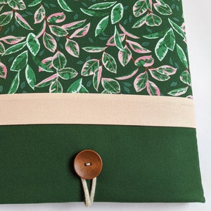 May include: A green canvas laptop sleeve with a pink and green floral pattern, a beige fabric band, and a wooden button closure.