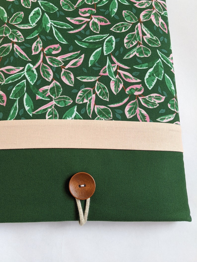 May include: A green canvas laptop sleeve with a pink and green floral pattern, a beige fabric band, and a wooden button closure.