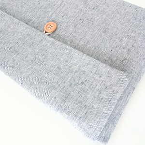 May include: Close-up of folded, light blue linen fabric with a visible weave. A natural wooden button is attached to the fabric with white thread. The fabric is folded over itself, creating a layered effect.