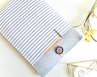 Puede incluir: Una funda para portátil de rayas grises y blancas con un botón marrón y un clip plateado. La funda está sobre una superficie blanca con un jarrón de flores amarillas y un soporte de metal dorado.