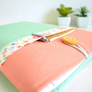 May include: A mint green and coral laptop sleeve with a floral patterned fabric detail and a brown button closure.