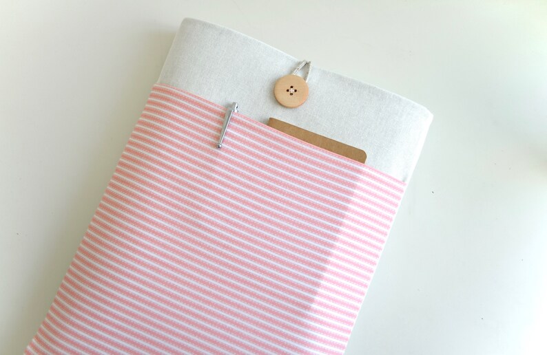 Op de afbeelding: Een roze en wit gestreepte laptophoes van stof met een witte klep van stof en een houten knoopsluiting.