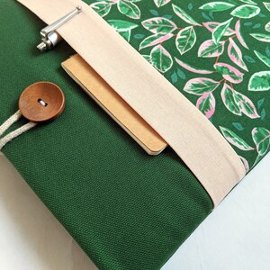 May include: A green canvas laptop sleeve with a pink and green floral pattern, a beige fabric flap, and a wooden button closure.