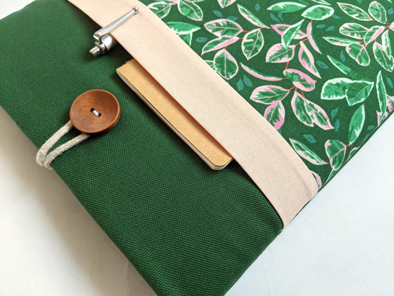 May include: A green canvas laptop sleeve with a pink and green floral pattern, a beige fabric flap, and a wooden button closure.