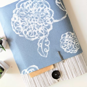 May include: A blue fabric book cover with a white floral pattern. The cover has a pocket on the front and a button closure.