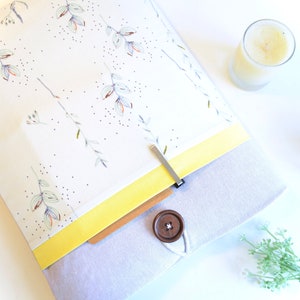 Puede incluir: Una funda para portátil gris y blanca con un patrón floral y un acento amarillo. La funda tiene una solapa de madera con un cierre de botón y un clip metálico.