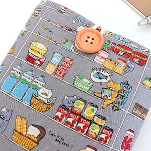 May include: A gray fabric book cover with a whimsical grocery store print featuring cats, food items, and shelves. The cover is secured with a large orange button. A pen and notebook are in the background.