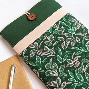 May include: A green laptop sleeve with a pink and green leaf pattern. The sleeve has a flap with a brown button and a pink fabric strip.