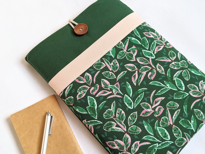 May include: A green laptop sleeve with a pink and green leaf pattern. The sleeve has a flap with a brown button and a pink fabric strip.