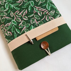 May include: A green laptop sleeve with a pink and green floral pattern on the inside. The sleeve has a flap with a light pink fabric strap and a brown button closure. A silver pen is tucked under the strap.