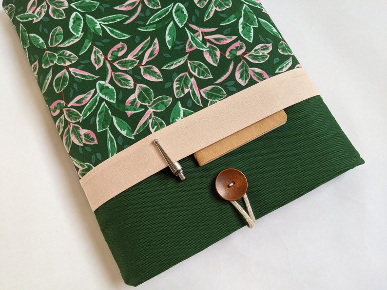May include: A green laptop sleeve with a pink and green floral pattern on the inside. The sleeve has a flap with a light pink fabric strap and a brown button closure. A silver pen is tucked under the strap.