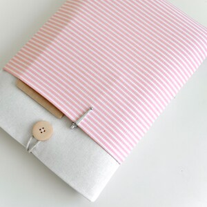 Op de afbeelding: Een roze en wit gestreepte laptophuls met een beige klep en een houten knoopsluiting. De hoes heeft een klein vakje aan de voorkant.