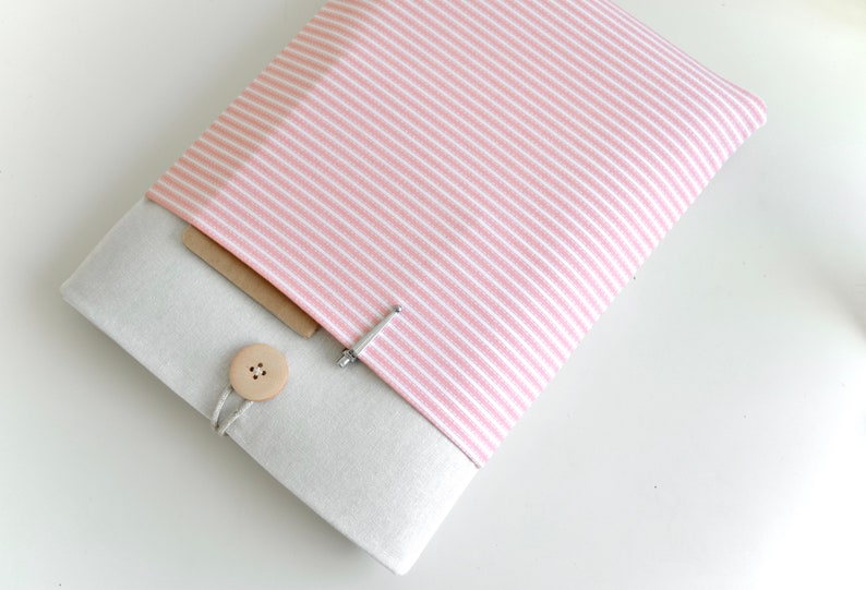 Op de afbeelding: Een roze en wit gestreepte laptophuls met een beige klep en een houten knoopsluiting. De hoes heeft een klein vakje aan de voorkant.