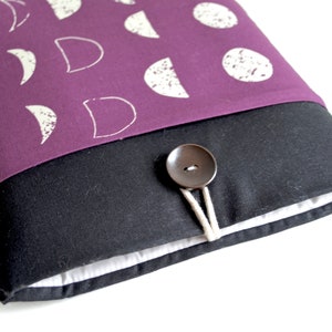 Può includere: Una custodia per laptop di colore viola con un motivo di lune e cerchi bianchi. La custodia ha una patta nera con una chiusura a bottone marrone e lacci in corda bianca.