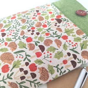 Puede incluir: Estuche rectangular de tela con estampado de bosque con erizos, manzanas, setas y hojas en tonos marrón, rojo y verde. Solapa de lino verde, asegurada con un botón y lazo de madera.
