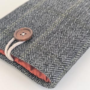 Puede incluir: Estuche gris y blanco con patrón de espiga y cierre de botón de madera. El forro interior es de color rojo anaranjado. El estuche probablemente está diseñado para guardar objetos pequeños o proteger un dispositivo.