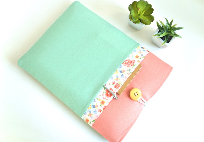 May include: A mint green laptop sleeve with a floral patterned pocket and a pink flap. The flap has a yellow button and a white string closure.