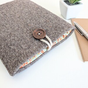 May include: A brown tweed fabric case with a wooden button fastening and a floral patterned lining. A notebook and pen are next to the case. The case is designed to hold a tablet or e-reader.