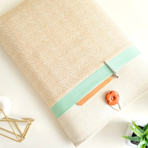 Puede incluir: Una funda para portátil beige con un patrón de espiga y una cinta verde menta. La funda tiene una solapa con un botón marrón y un clip plateado.