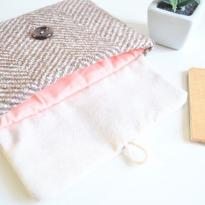 Pode incluir: Uma carteira de mão de tweed marrom e branco com forro rosa e fecho de botão marrom. A carteira de mão está aberta e o forro é visível. A carteira de mão está sobre uma superfície branca.