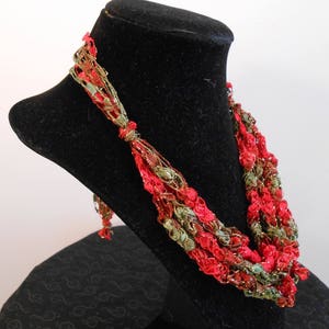 May include: A multi-strand crocheted necklace with red, green, and gold yarn. The necklace is displayed on a black mannequin bust.