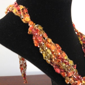 May include: A multi-strand crocheted necklace with a mix of red, orange, and gold yarn. The necklace is displayed on a black velvet bust form.